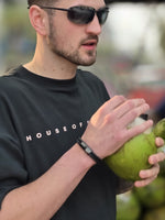 Load image into Gallery viewer, Model wearing sunglasses and a hunter green 'HOUSE OF SWASHA' t-shirt holding a coconut.