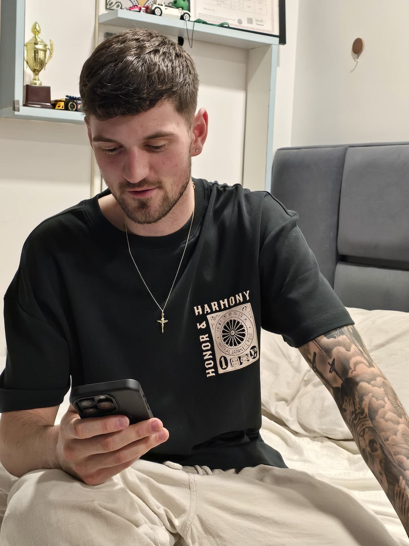 Model sporting the House of SwaSha Honor and Harmony t-shirt, sitting on a bed using a phone. Casual comfort, sustainable fashion.
