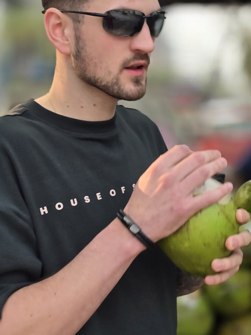 Model wearing sunglasses and a hunter green 'HOUSE OF SWASHA' t-shirt holding a coconut.