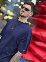 Load image into Gallery viewer, Model wearing a navy blue t-shirt with sunglasses and a pendant, standing in front of a red sculpture.